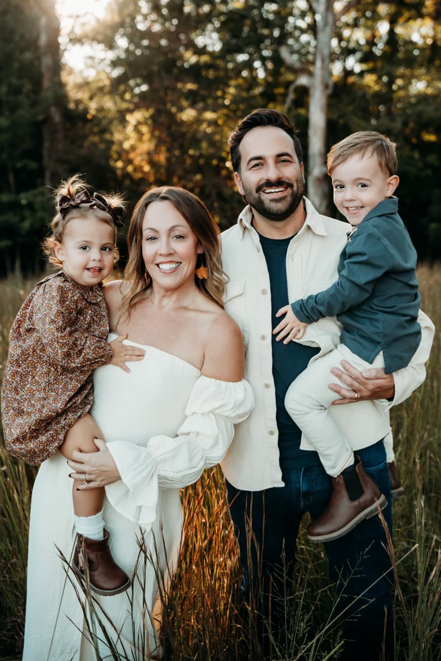 Our family: two parents, each holding one of our two toddlers, smiling in a field of tall grass
